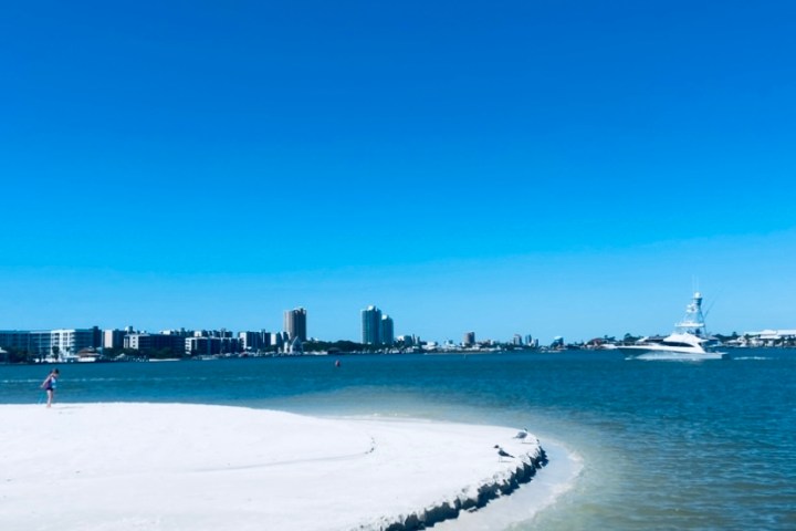 Sandy beach, person walking, boat on water, city skyline in background under clear blue sky.