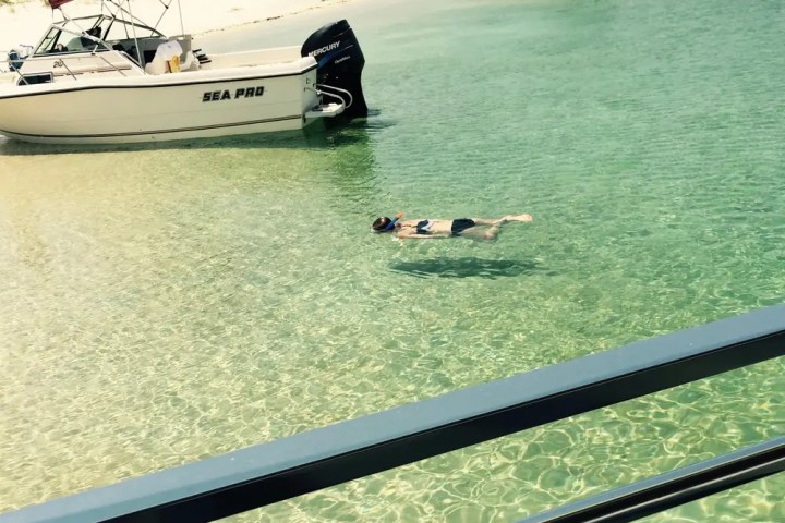 Person snorkeling near anchored boats in clear, shallow water by a sandy beach.