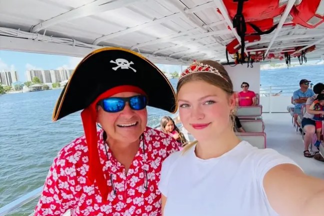 Man in pirate hat and woman in tiara take a selfie on a boat with others seated in the background.